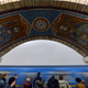 Mosaic tiles in red blue and y ellow form a design over an archway in a subway station. Below the arch are passengers waiting for a blue subway train.